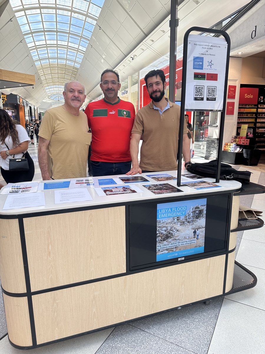 Hamza Katranji in Morocco’s red football shirt, standing between two Libyan members of the Jeeran community behind the club’s UNICEF stall at Pacific Epping, with the Libya Flood Emergency poster on the front.